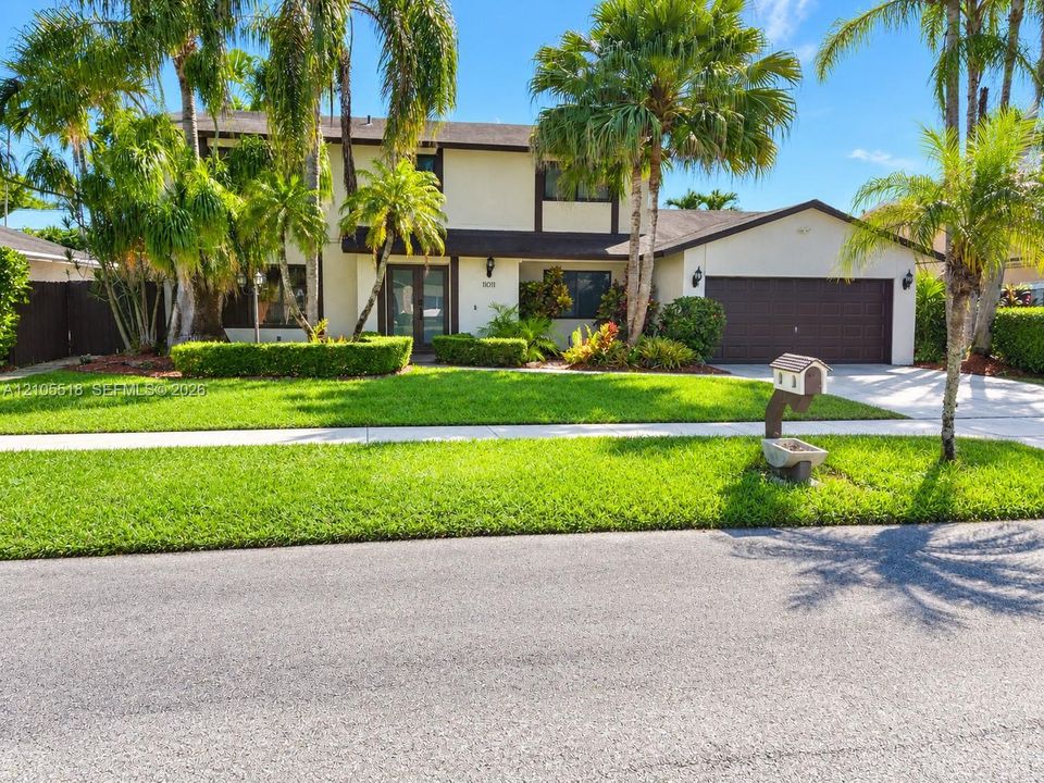 Two-story exterior featuring a stucco facade, dark-toned trim, a multi-car garage, manicured landscaping, and mature palm trees