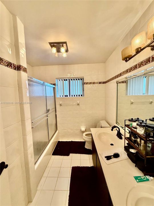 Full bathroom featuring white tile wall surrounds with decorative mosaic accent banding, a dual-sink vanity with integrated countertop, a sliding glass door shower enclosure, and multi-bulb wall-mounted lighting