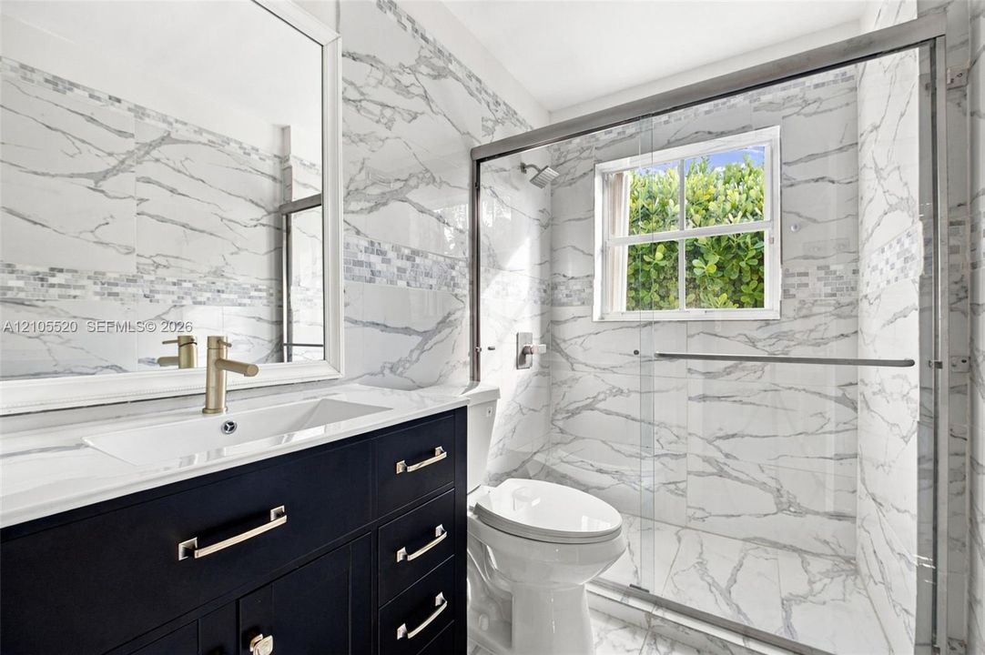 Dark navy vanity cabinet with metallic hardware, white countertop, large-format veined tile wall surfacing, glass-enclosed shower, and framed vanity mirror