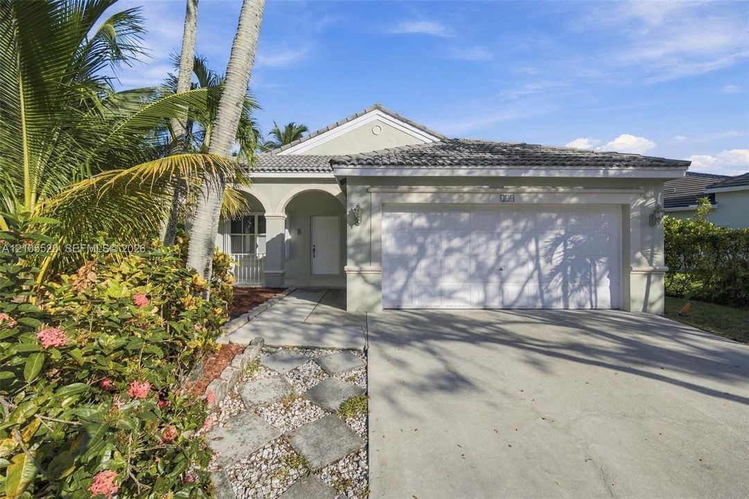 Single-story exterior featuring a concrete tile roof, arched entryway, and two-car garage, complemented by a stone paver walkway and established tropical landscaping
