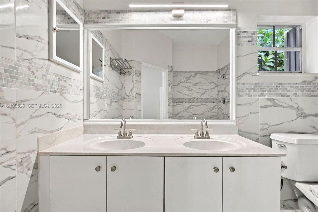 Double vanity featuring a stone countertop, dual undermount sinks, large rectangular mirror, and full-height stone-finish wall tile with accent inlay