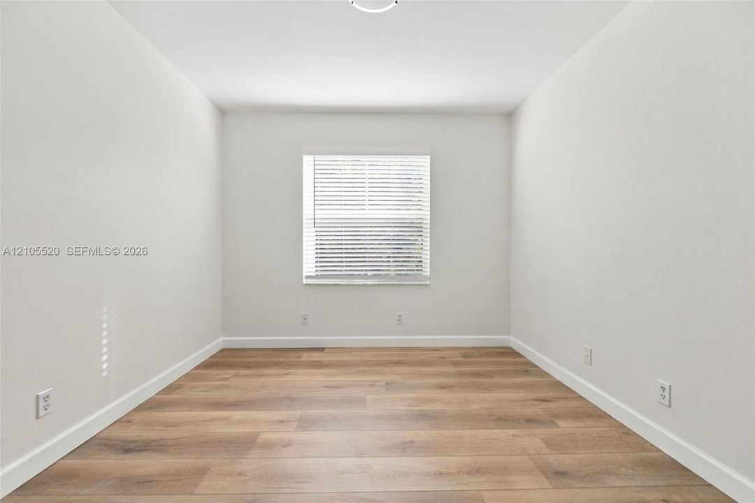 Neutral interior space featuring wood-finish flooring, crisp white walls, and a single window with horizontal blinds