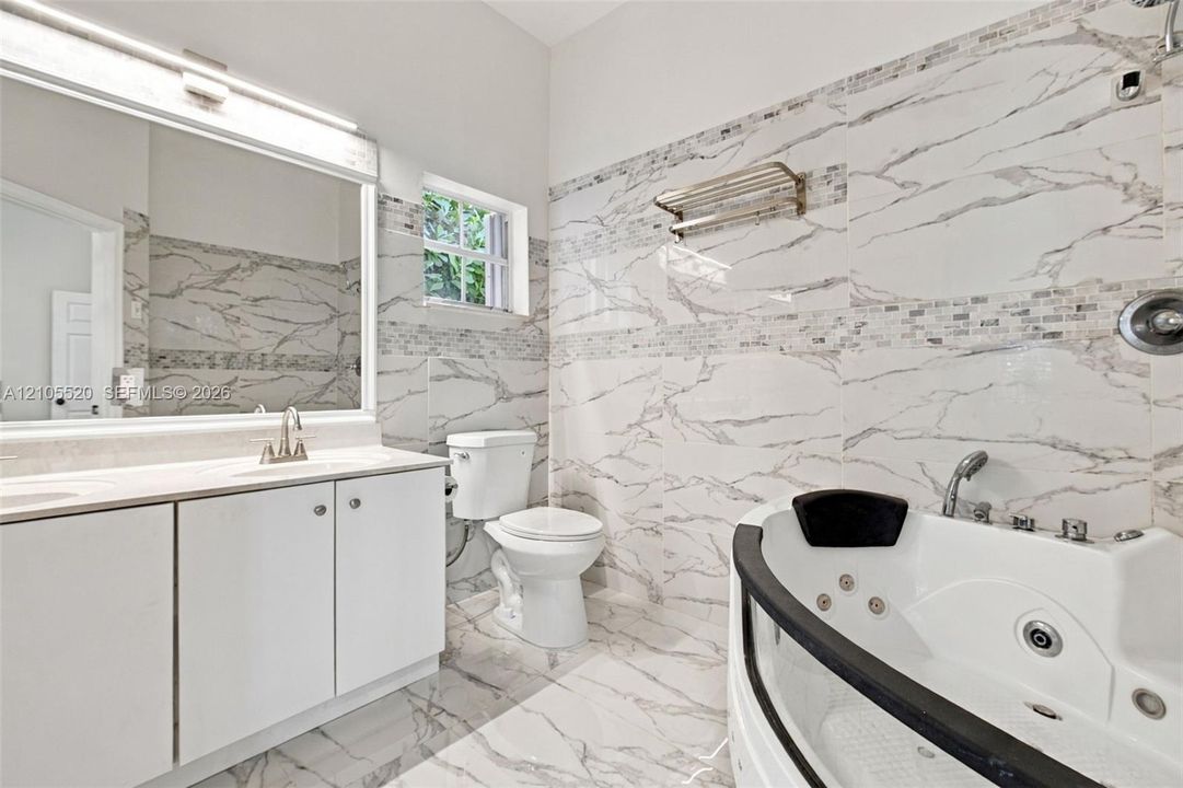Bathroom featuring floor-to-ceiling large-format tile with stone-pattern finish, jetted soaking tub, white vanity cabinet with countertop basin, and oversized wall-mounted mirror