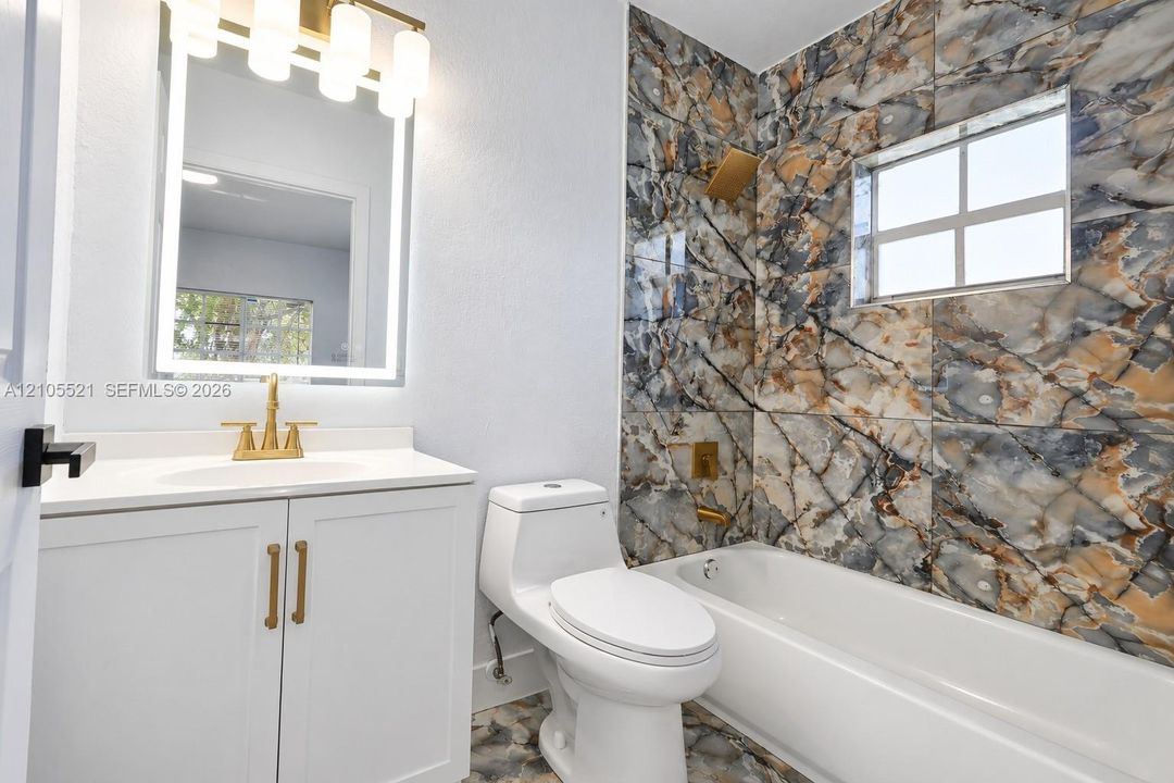 White vanity with a solid surface countertop, illuminated mirror, and a gold-tone faucet