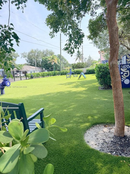 Expansive green space featuring artificial turf, mature trees, a decorative bench, and a privacy hedge