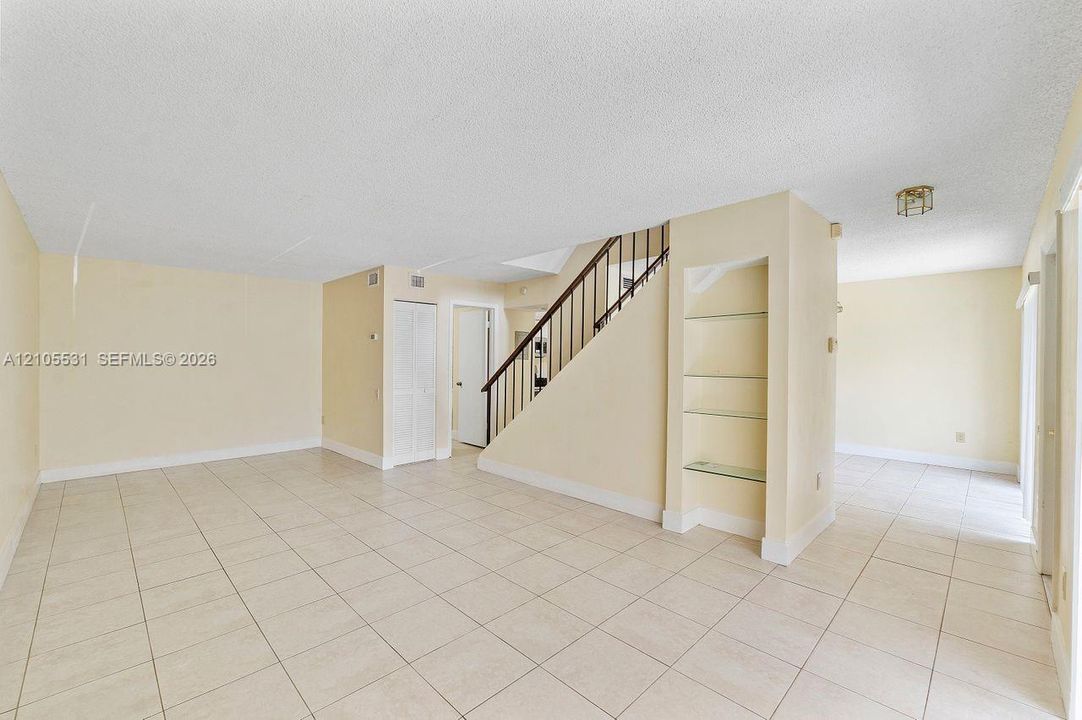 Spacious room featuring neutral tile flooring, a staircase with dark wood railings, and built-in shelving with glass inserts