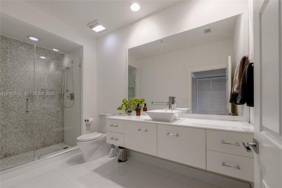 Floating white vanity with a rectangular vessel sink, solid-surface countertop, frameless glass shower enclosure, and textured stone-finish shower wall