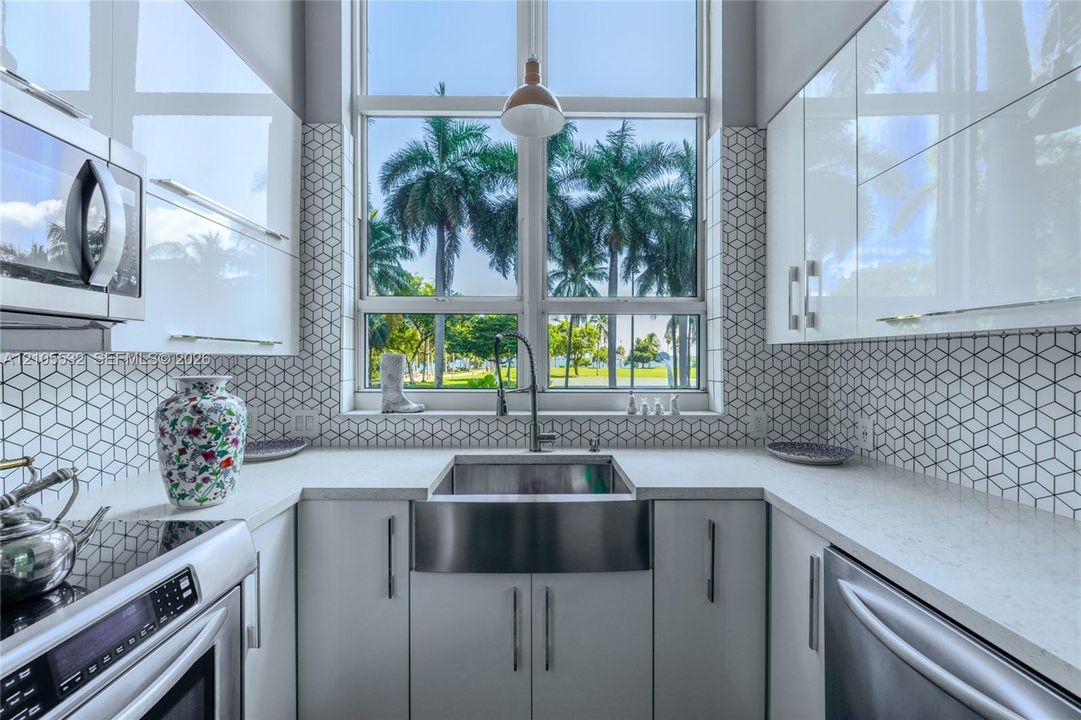 Kitchen featuring high-gloss cabinetry, stone-finish countertops, stainless steel farmhouse sink, and geometric-patterned backsplash tile, accented by a floor-to-ceiling window