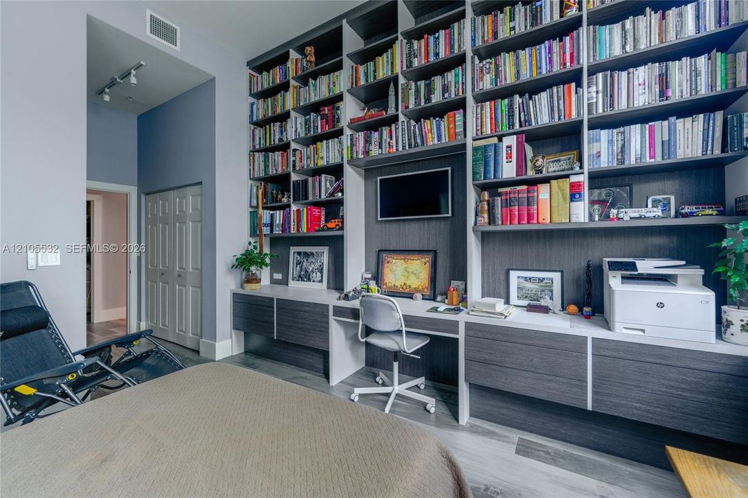 Floor-to-ceiling custom built-in shelving, integrated desk workspace, dual-tone cabinetry with wood-finish detailing, track lighting, and wood-finish flooring