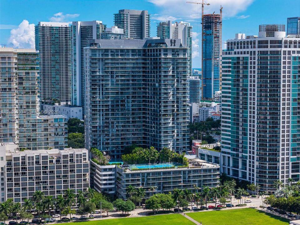 High-rise residential building featuring extensive glass balcony railings, a rooftop pool, a tennis court, and mature landscaping