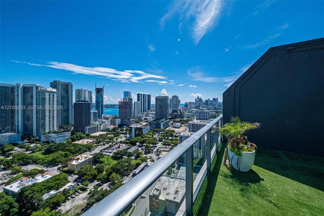 Balcony featuring synthetic turf flooring, metal and glass railing, and urban skyline backdrop
