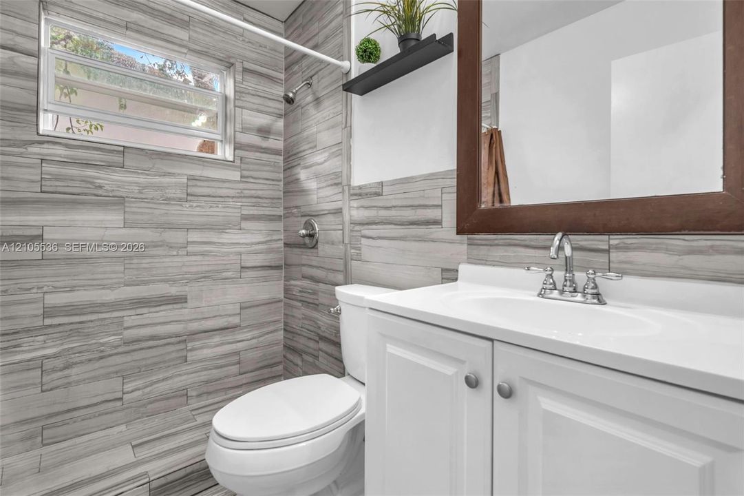 Bathroom featuring horizontally-laid tile wall treatments, white cabinetry with an integrated basin, a polished chrome double-handle faucet, and an awning-style window