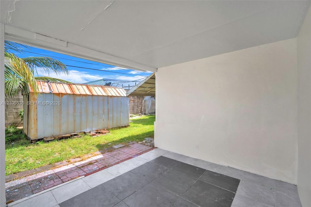 Covered patio with large-format tile flooring, open-air design, and direct access to a fenced rear yard featuring a grass lawn and detached metal utility structure