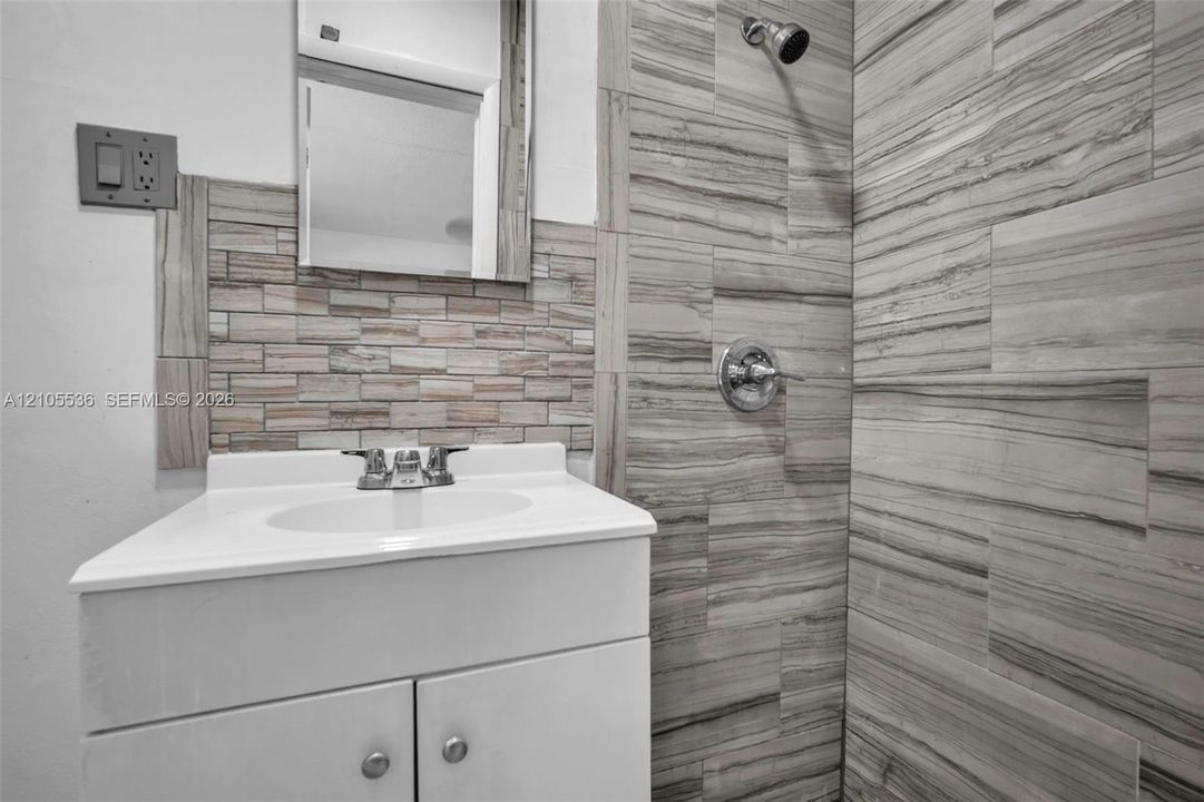 Bathroom vanity featuring a white cabinet, integrated sink, chrome faucet, and mosaic tile backsplash, accompanied by a shower enclosure finished in large-format linear-patterned tile with chrome fixtures