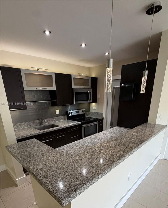 Kitchen featuring stone countertops, modern cabinetry with glass-panel accents, stainless steel appliances, tile backsplash, and contemporary pendant lighting
