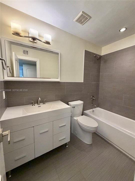 White vanity with rectangular integrated sink, three-light wall-mounted fixture, large framed mirror, and floor-to-ceiling gray tiled shower surround