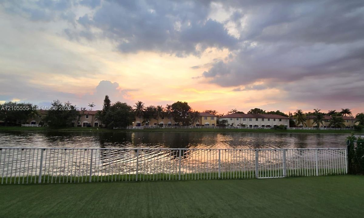 Waterfront property featuring a manicured lawn, a white metal perimeter fence, and a tranquil lake backdrop