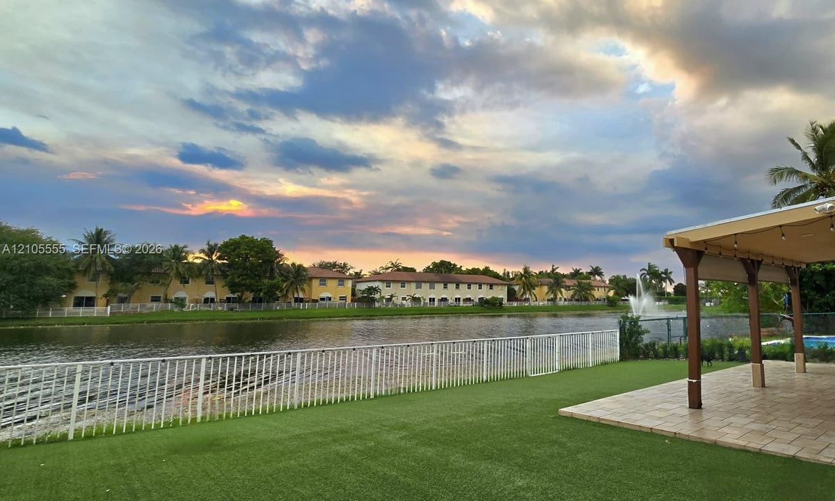 Waterfront grounds featuring a covered paver patio, synthetic turf lawn, metal perimeter fencing, and a decorative water fountain