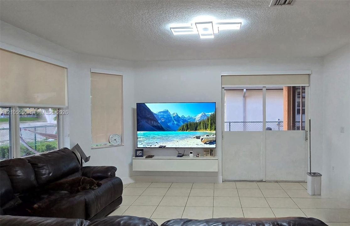Living area featuring large format tile flooring, a geometric LED ceiling fixture, wall-mounted floating cabinetry, and sliding glass doors with roller shades