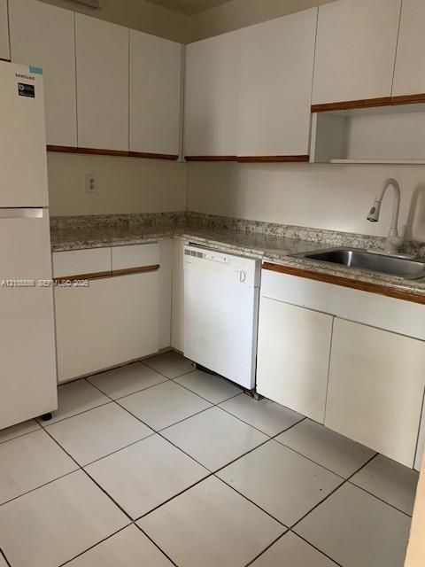 White cabinetry with wood-finish trim, speckled countertops, stainless steel sink, and white tile flooring