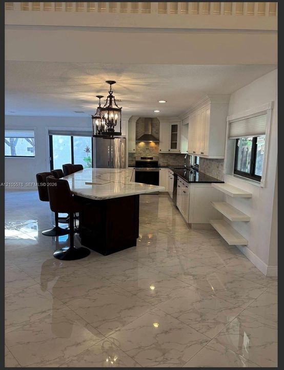 Kitchen featuring a large central island with light-toned stone-finish countertop, white cabinetry, dark stone-finish perimeter counters, polished tile flooring, and decorative hanging pendant lighting
