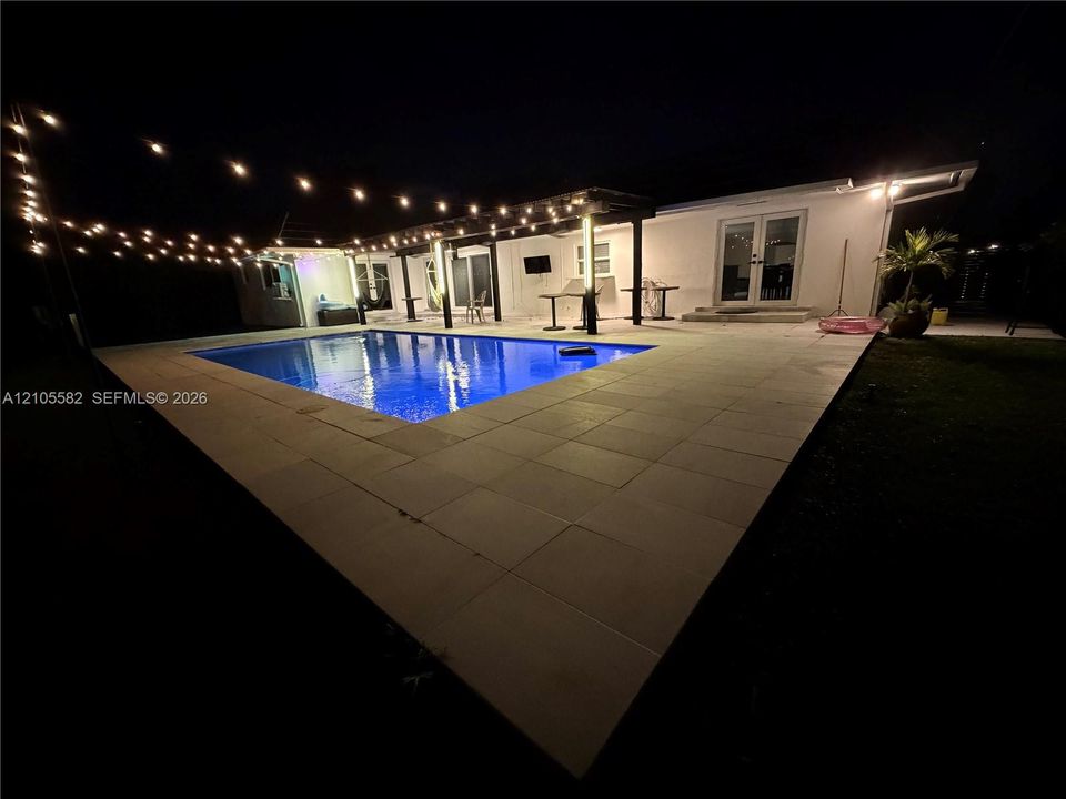 In-ground pool surrounded by a large paver patio, featuring string lighting and a covered pergola