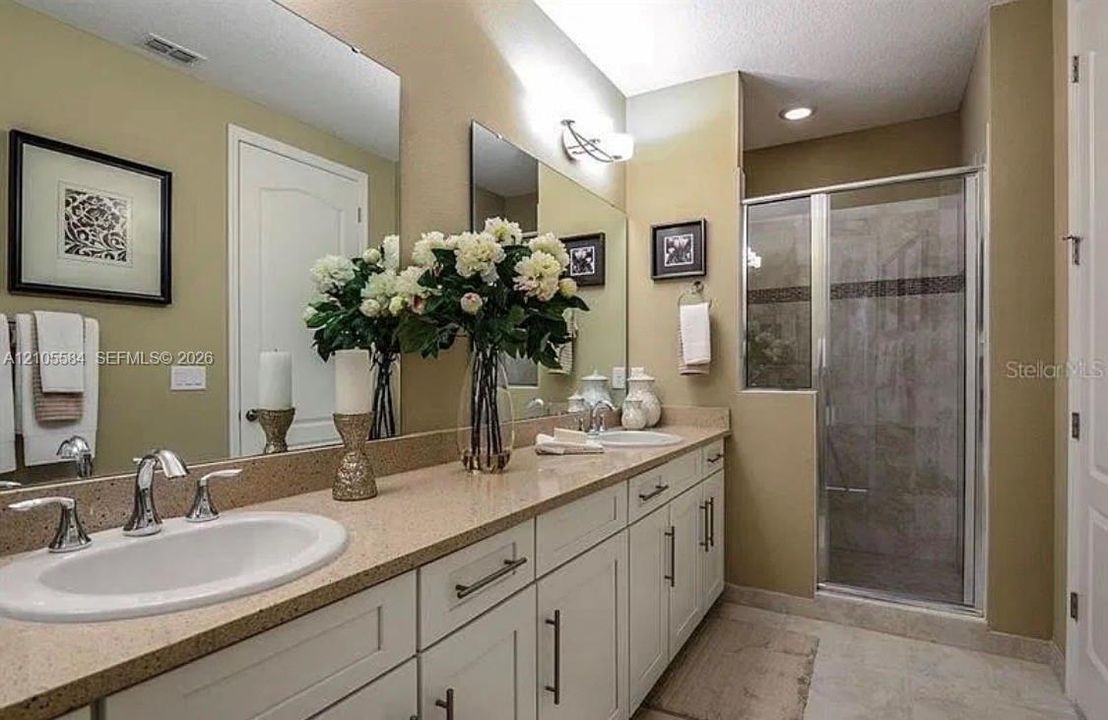 Dual-sink vanity with light-tone countertops, white cabinetry with silver-tone hardware, large wall-mounted mirrors, and a glass-enclosed shower with tiled surround