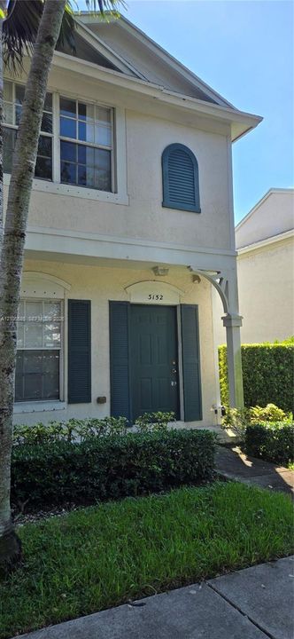 Two-story exterior featuring a light-toned stucco facade, decorative arched shutter detail, covered entryway with corbel supports, and multi-pane windows