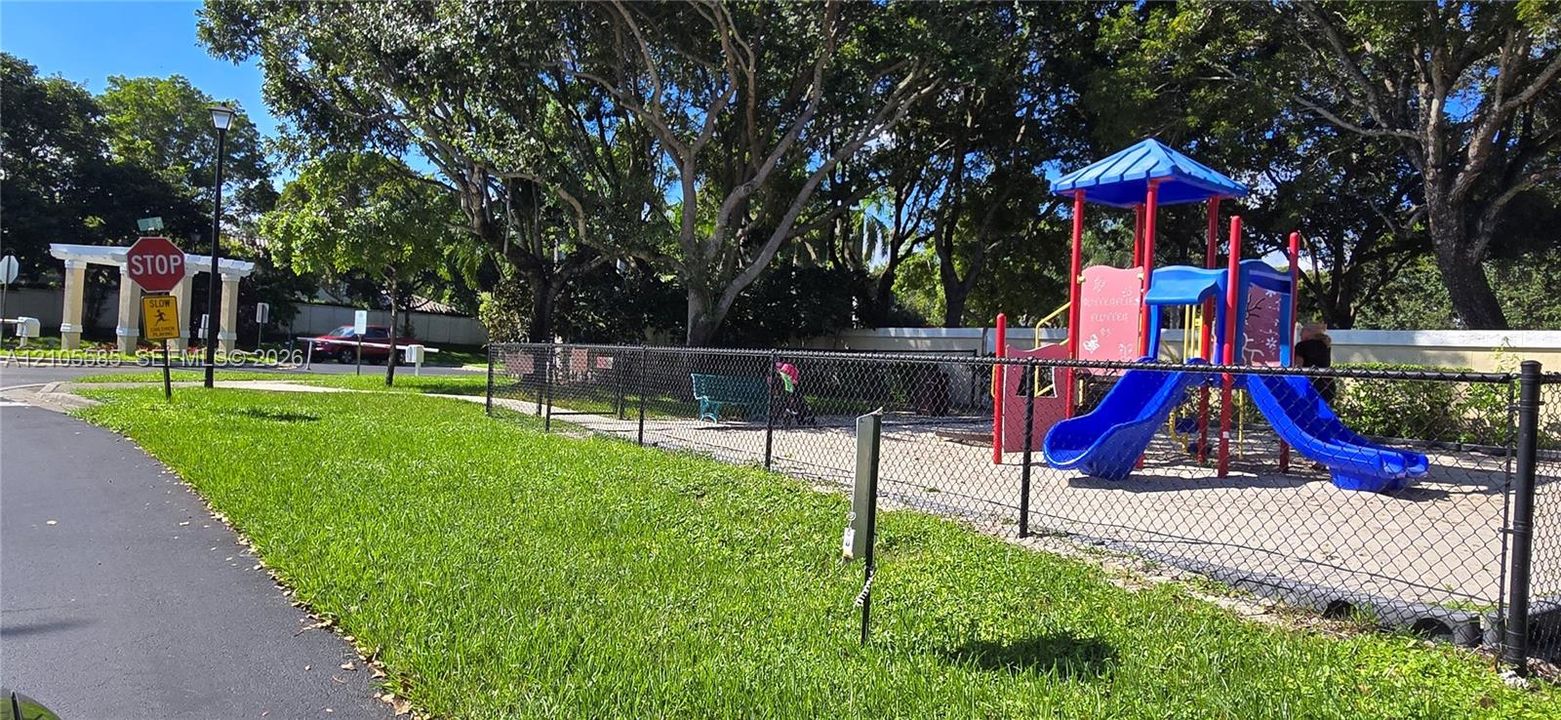 Recreational area featuring a fenced playground, paved walkways, mature trees, and a shaded structure