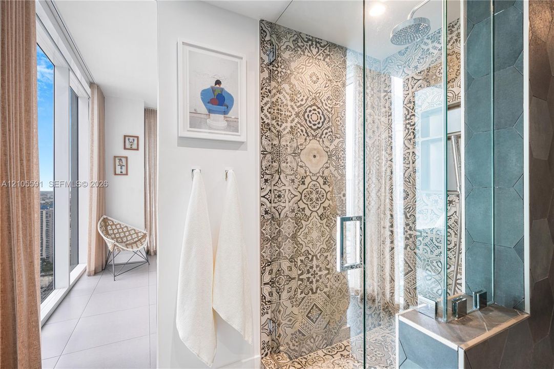 Full-height window with city backdrop, glass shower enclosure, decorative patterned wall tile, rainfall showerhead, and a built-in shower niche
