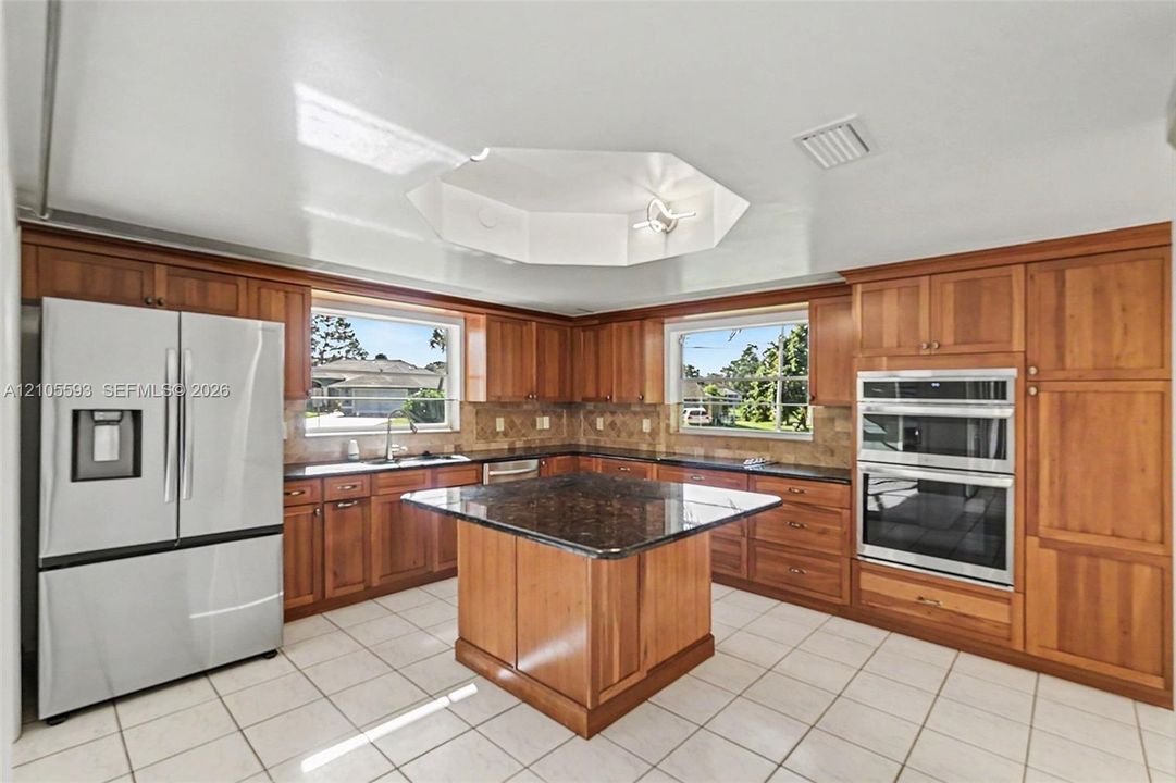 Kitchen featuring natural wood cabinetry, stone-surfaced central island, stainless steel double wall ovens, tile flooring, and a recessed hexagonal ceiling detail