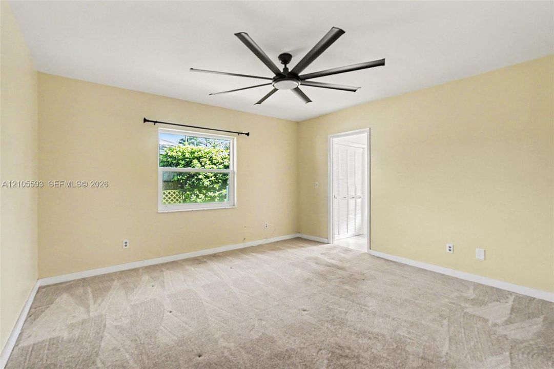 Neutral-toned interior room featuring wall-to-wall carpeting, an oversized multi-blade ceiling fan, and a single-pane window with a curtain rod
