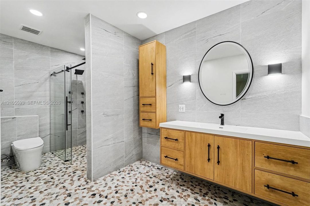 Wall-mounted wood-finish vanity with a rectangular sink, walk-in glass shower enclosure, stone-tiled walls, integrated cabinetry, and round vanity mirror