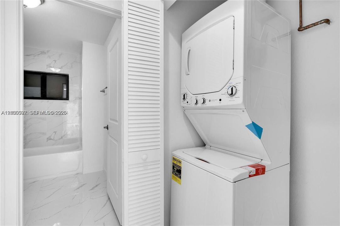 Laundry area featuring a stackable washer and dryer unit, louvered closet door, and stone-finish flooring extending into the bathroom with a shower-tub combination and wall tiling