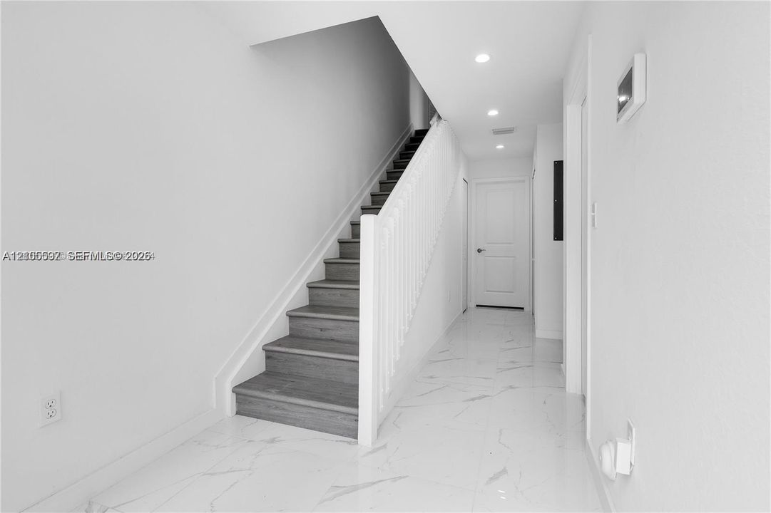 Interior hallway featuring polished stone-finish tile flooring, recessed ceiling lighting, and a staircase with wood-finish treads and a painted white balustrade
