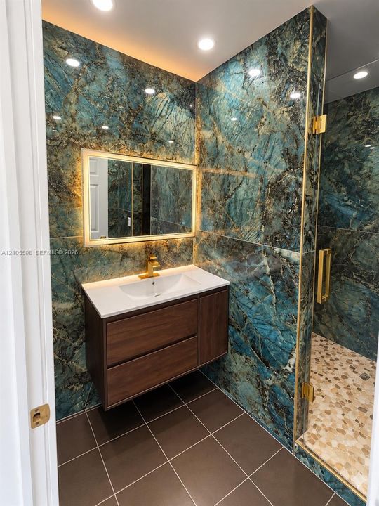 Floating vanity with an integrated sink, illuminated mirror, floor-to-ceiling accent wall, gold-toned fixtures, and a shower enclosure featuring mosaic tile