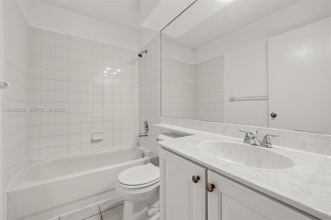 Bathroom featuring a white bathtub with a tile surround, vanity with a light-toned stone-patterned countertop, integrated oval sink, and a full-width wall mirror
