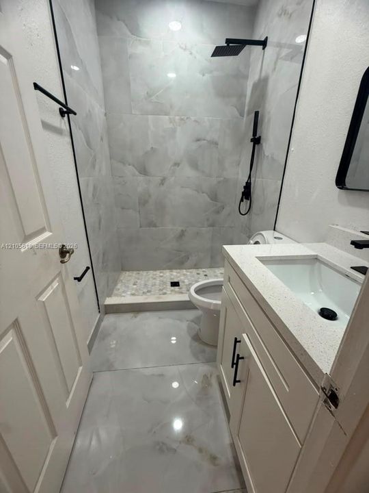 Bathroom featuring large-format tile flooring, a walk-in shower with floor-to-ceiling tile surround and a pebble-tiled base, a vanity with a quartz countertop, and matte black hardware