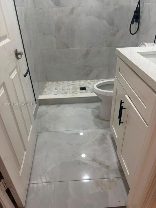 Bathroom featuring large-format porcelain tile flooring, a shower with floor-to-ceiling tile surround and a stone-pebble shower floor, quartz vanity countertop, and matte black cabinet hardware