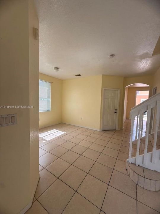 Interior space featuring neutral-toned tile flooring, white stairwell with balustrade, arched architectural detail, multi-panel door, and window with blinds