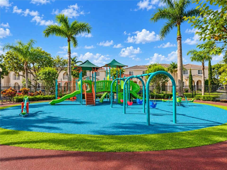 Playground facility featuring multiple slides, swing sets, and climbing structures on a multi-colored rubberized surface, surrounded by manicured landscaping and palm trees