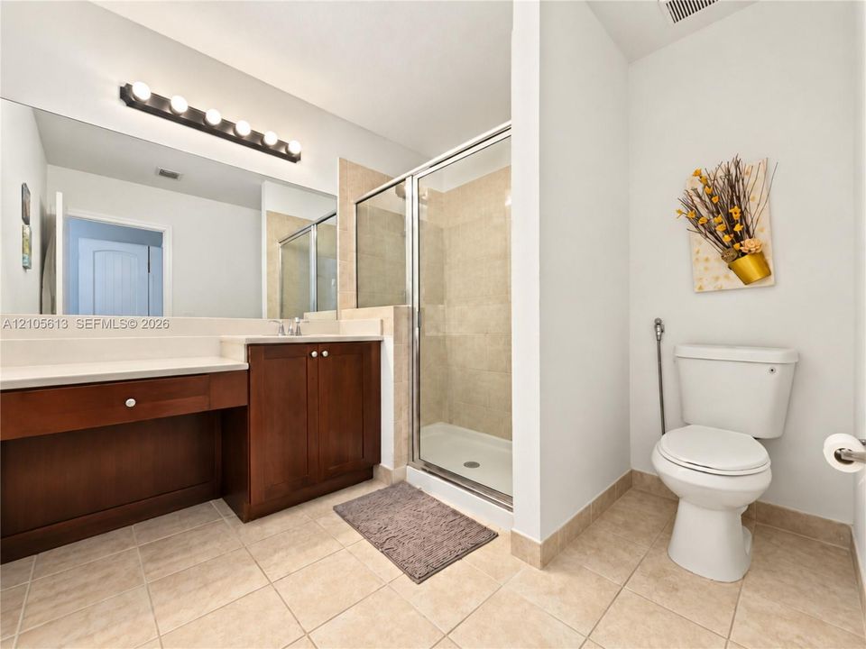 Bathroom featuring a wood-finish vanity with a light-toned countertop, a large wall-mounted mirror with multi-bulb light fixture, a glass-enclosed shower with tile surround, and tile flooring