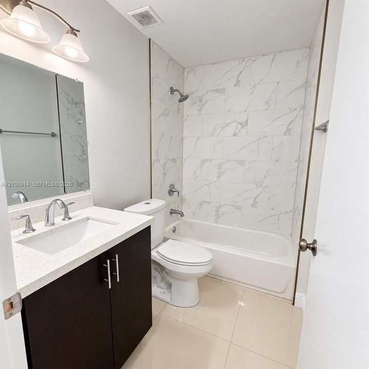 Bathroom featuring a vanity with a light-toned solid surface countertop, dark-finish cabinetry, a white porcelain tub with floor-to-ceiling surround tile, and polished chrome hardware