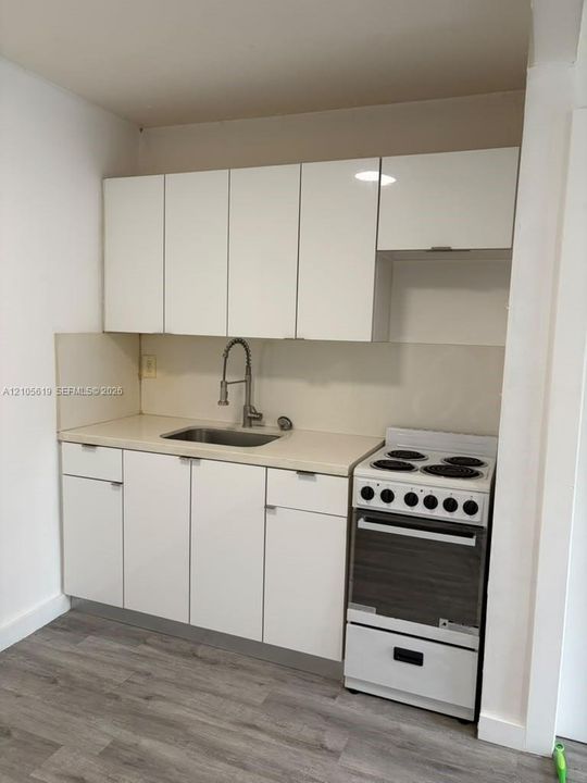 Kitchenette featuring high-gloss white flat-panel cabinetry, solid-surface countertop, stainless steel drop-in sink with a high-arch gooseneck faucet, four-burner electric range, and wood-finish flooring