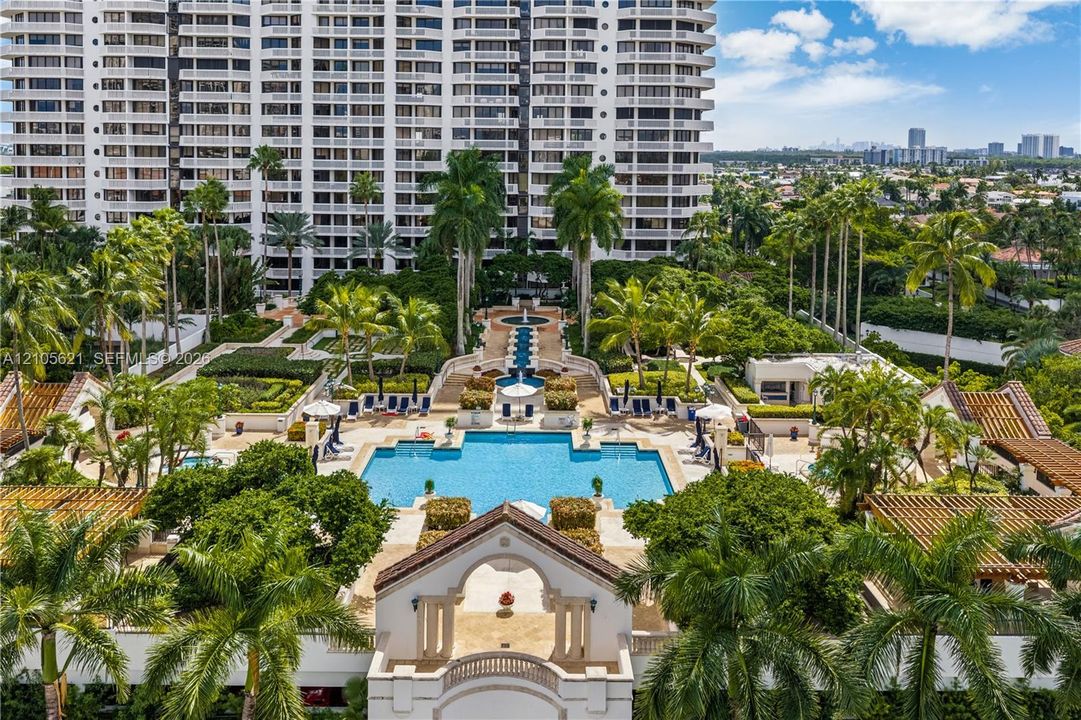 High-rise residential building featuring a large swimming pool, tiered fountain water feature, expansive stone-paved sun deck, and mature palm tree landscaping
