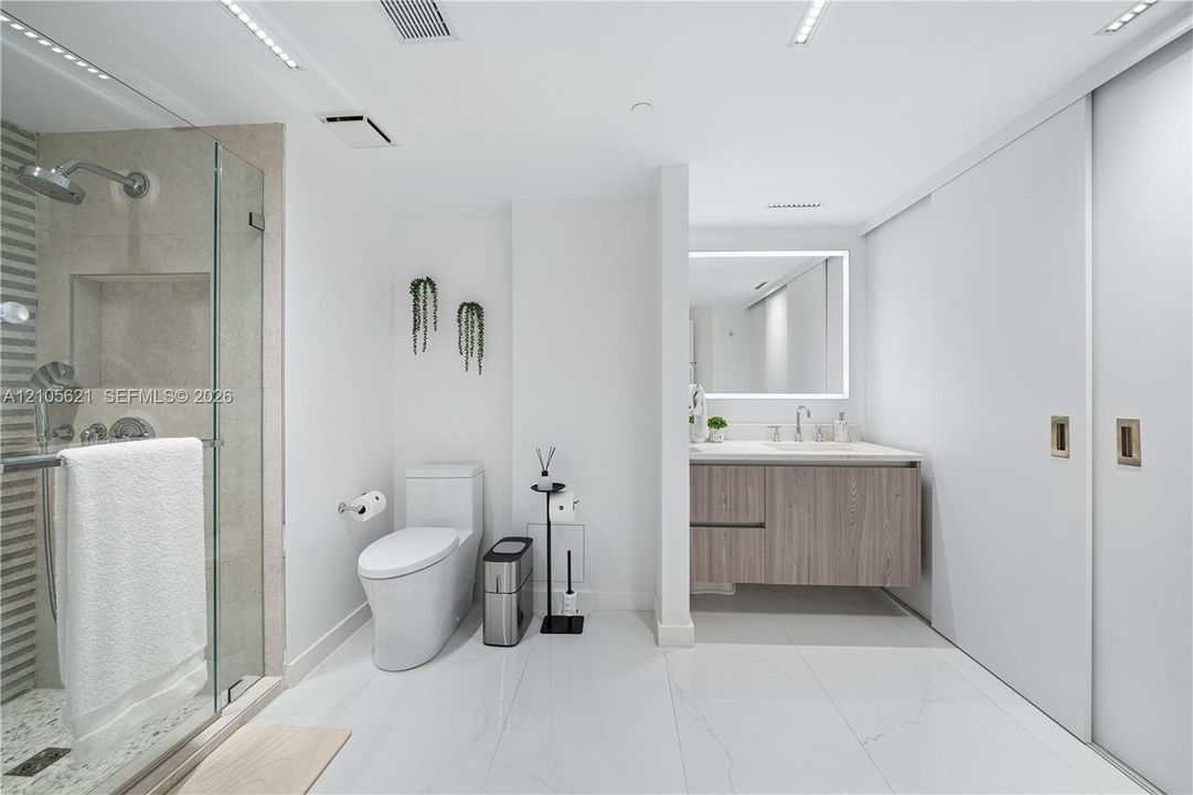 Modern bathroom featuring a glass-enclosed shower, wall-mounted wood-finish vanity with a light countertop, LED-backlit mirror, and white tile flooring