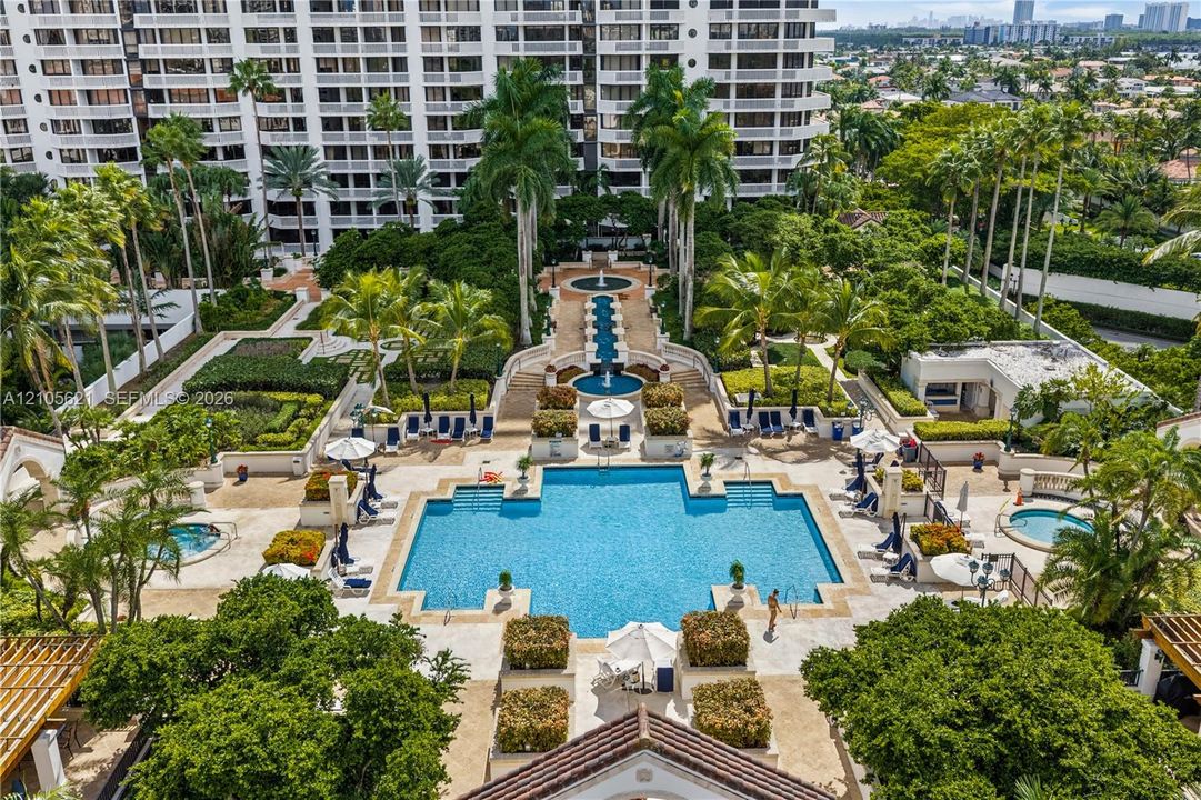 Resort-style pool area featuring a multi-tiered fountain, stone-paved sun deck, mature palm trees, and two separate hot tubs