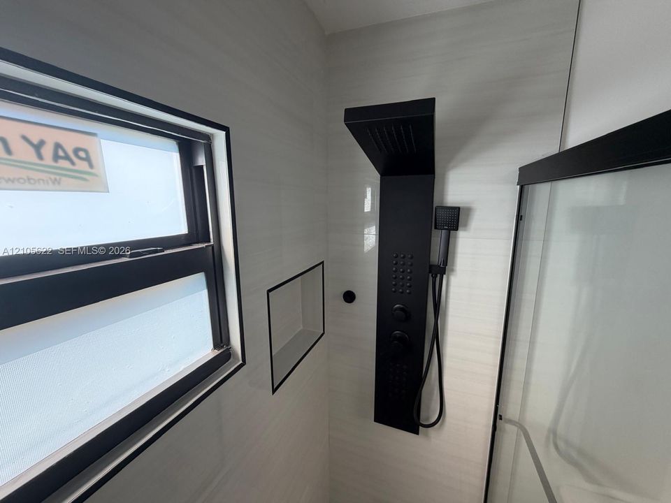 Walk-in shower featuring light-toned tile walls, a matte black multi-function shower panel with an integrated handheld sprayer, a built-in recessed wall niche, and a window with a matte black frame