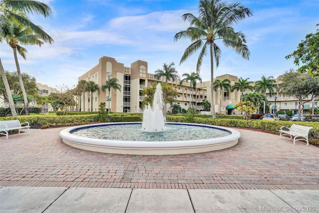 Circular stone fountain centerpiece set within a brick paver courtyard, surrounded by manicured landscaping, mature palm trees, and structural stucco exterior