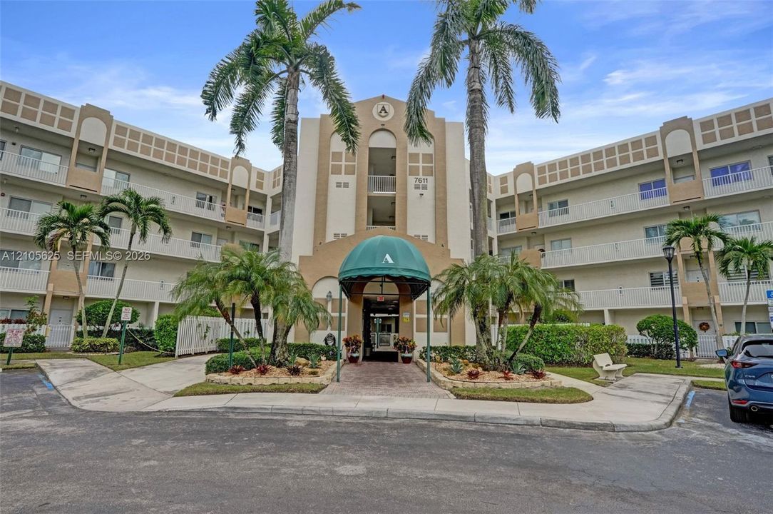 Multi-story exterior featuring a central arched entryway with a green metal awning, brick paver accents, and mature palm landscaping